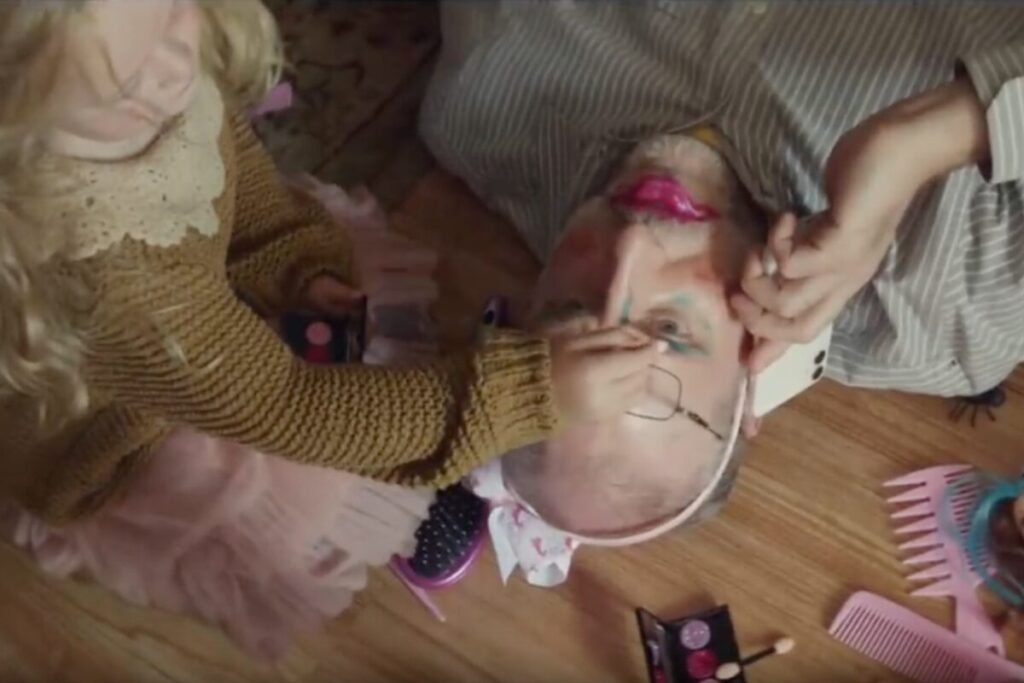 Parent lying on the floor while child applies makeup, highlighting the humor of parenting during remote work in Apple's ad.